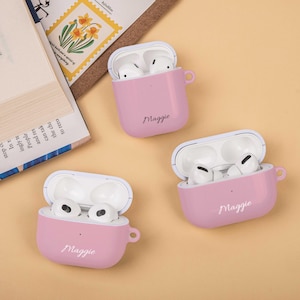 May include: Three pink AirPod cases, each with the name "Maggie" in cursive. The cases are open, revealing white AirPods. The cases have a small loop on the side. A book and a floral stamp are in the background.