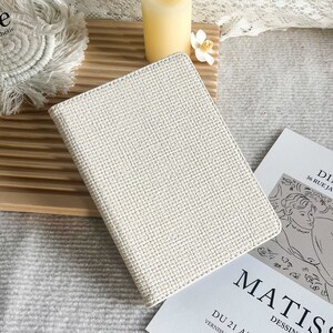 May include: A white woven notebook with a textured cover. The notebook is sitting on a wooden surface with a white candle and a white flower.