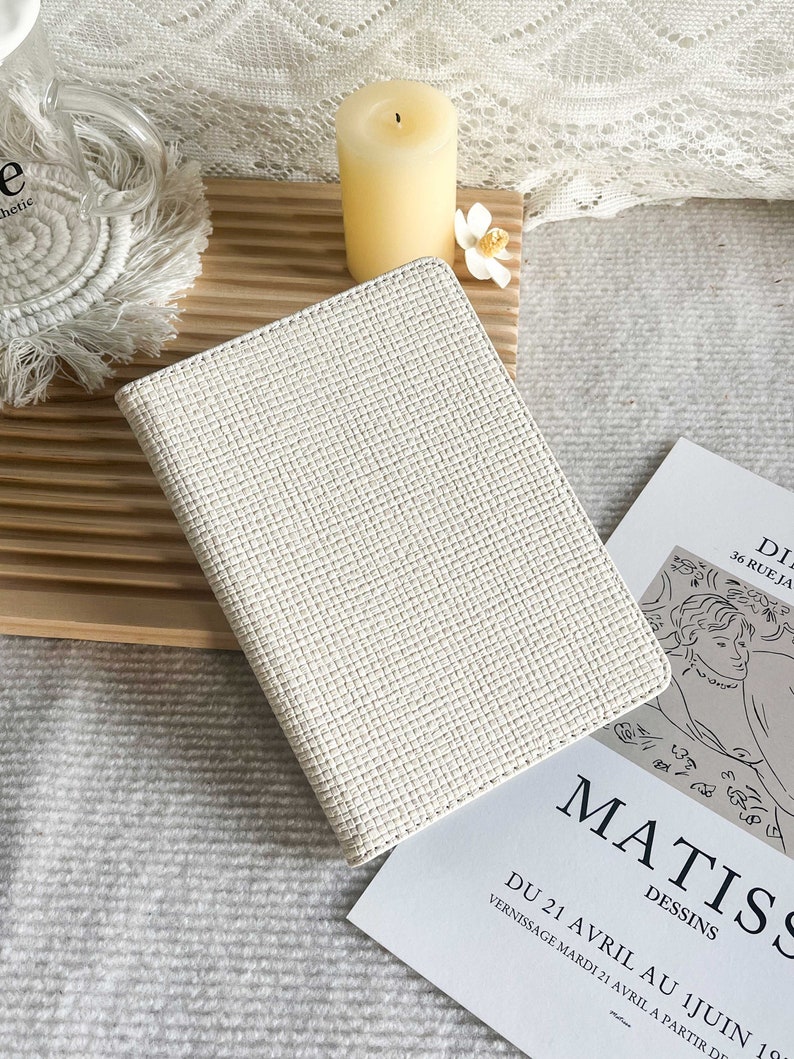 May include: A white woven notebook with a textured cover. The notebook is sitting on a wooden surface with a white candle and a white flower.