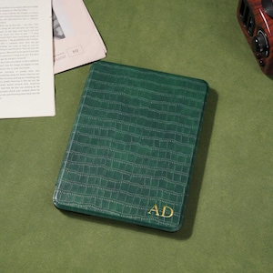 May include: A green, rectangular journal with a crocodile skin texture and gold-colored initials "AD". The journal is on a green surface, with a newspaper and a vintage-style radio in the background.