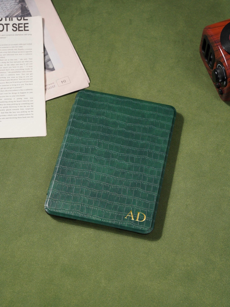May include: A green, rectangular journal with a crocodile skin texture and gold-colored initials "AD". The journal is on a green surface, with a newspaper and a vintage-style radio in the background.