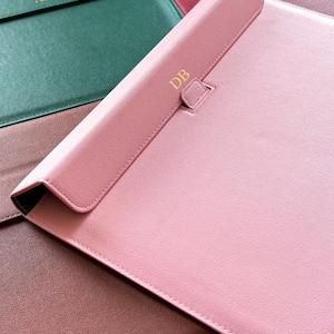 May include: A collection of personalized leather laptop sleeves in various colors, including pink, green, and burgundy. Each sleeve features gold-colored initials, such as "AD" and "DB", adding a custom touch. The sleeves have a flap closure.