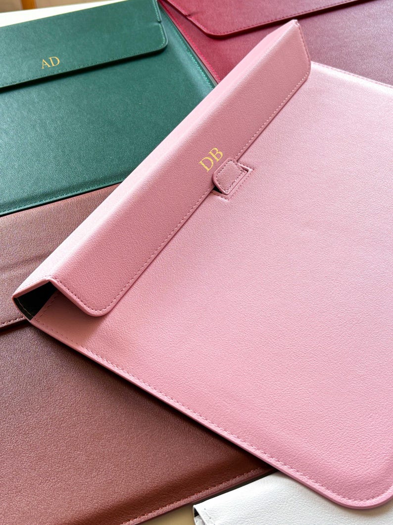 May include: A collection of personalized leather laptop sleeves in various colors, including pink, green, and burgundy. Each sleeve features gold-colored initials, such as "AD" and "DB", adding a custom touch. The sleeves have a flap closure.