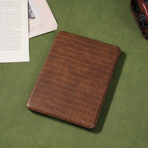 May include: A brown, rectangular tablet case with a textured, crocodile-skin pattern. The case is on a green surface, with a newspaper and a radio in the background.