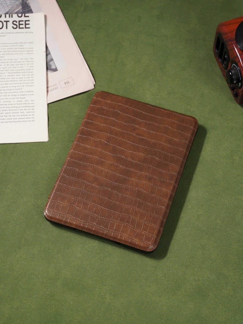 May include: A brown, rectangular tablet case with a textured, crocodile-skin pattern. The case is on a green surface, with a newspaper and a radio in the background.