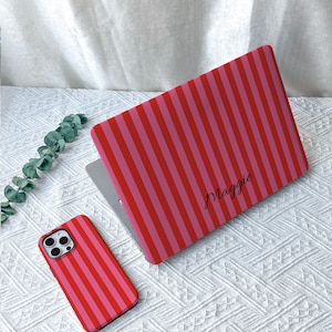 May include: A laptop cover and a matching smartphone case, both featuring a vibrant pattern of vertical red and pink stripes. The laptop cover displays the name Maggie in a black cursive script font, while the smartphone case shows the same striped design.