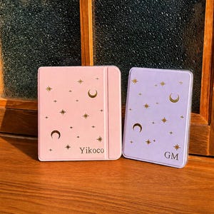 May include: A light pink e-reader case with gold stars, moons, and the text Yikoco, alongside a light purple e-reader case with gold stars, moons, and the text GM. Both cases feature a smooth finish and a celestial pattern on the front covers.