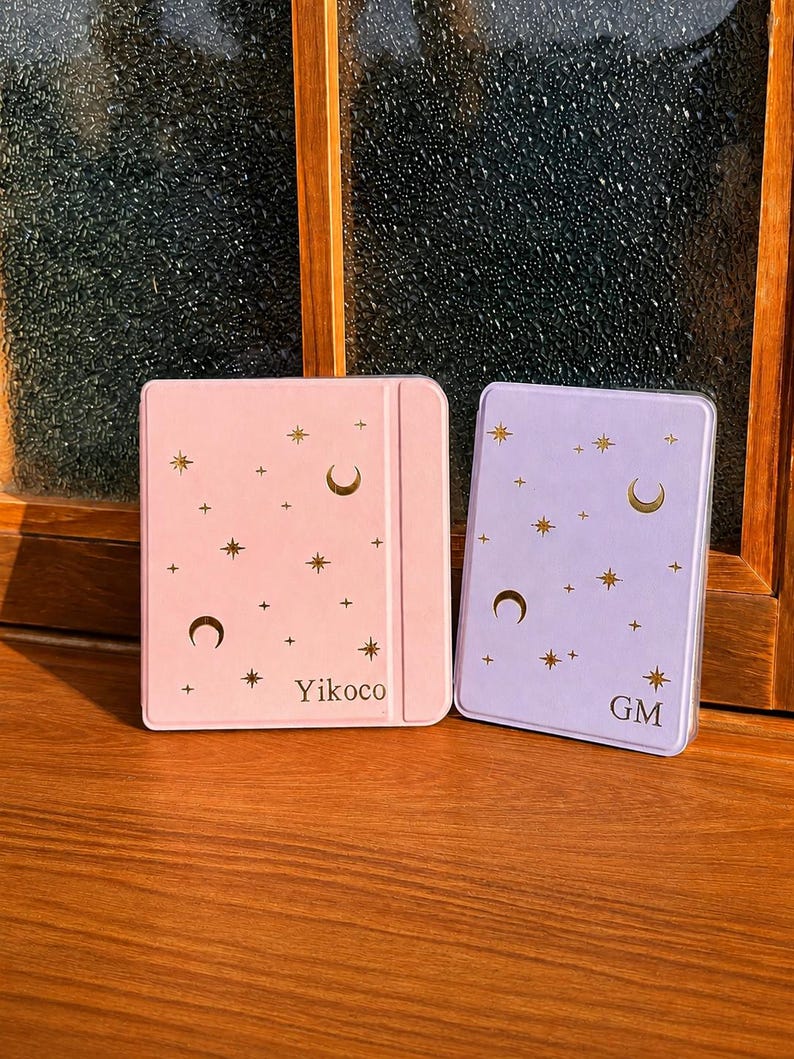 May include: A light pink e-reader case with gold stars, moons, and the text Yikoco, alongside a light purple e-reader case with gold stars, moons, and the text GM. Both cases feature a smooth finish and a celestial pattern on the front covers.