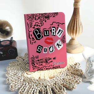 May include: A pink notebook featuring the words "BURN BOOK" in black and white, a red lipstick mark, and doodles. It rests on a woven placemat, with a decorative candle holder and a gramophone visible in the background.