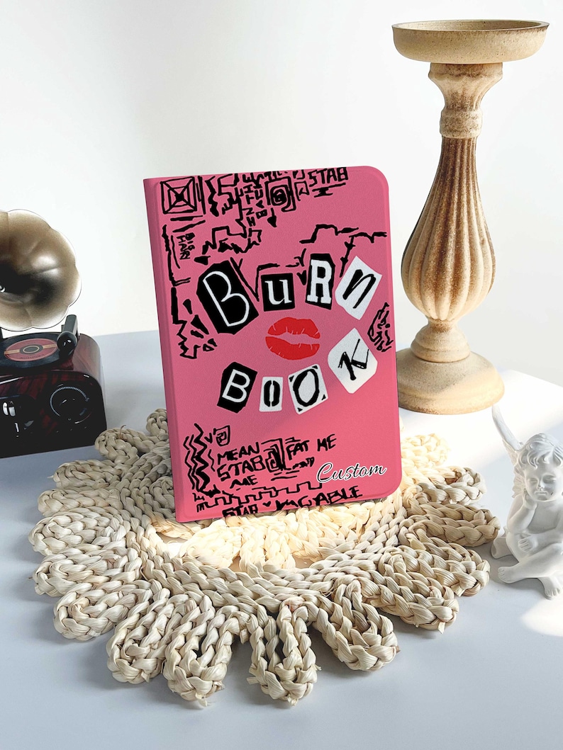 May include: A pink notebook with the words "BURN BOOK" in black and white lettering, a red lipstick kiss, and various doodles. The notebook is on a woven placemat with a decorative candle holder and a small gramophone in the background.