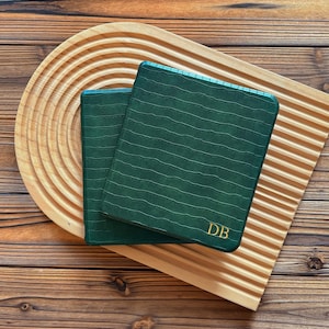 May include: Two dark green, textured, square notebooks with gold initials "DB" are stacked on a wooden tray with a curved design. The tray and the surface beneath are made of wood.