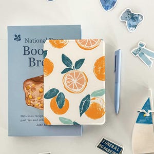 May include: A white notebook with a repeating pattern of orange slices and green leaves. The notebook is closed and lying on a white surface. There are several blue stickers around the notebook, including a clock, a teddy bear, a box, and a bow.