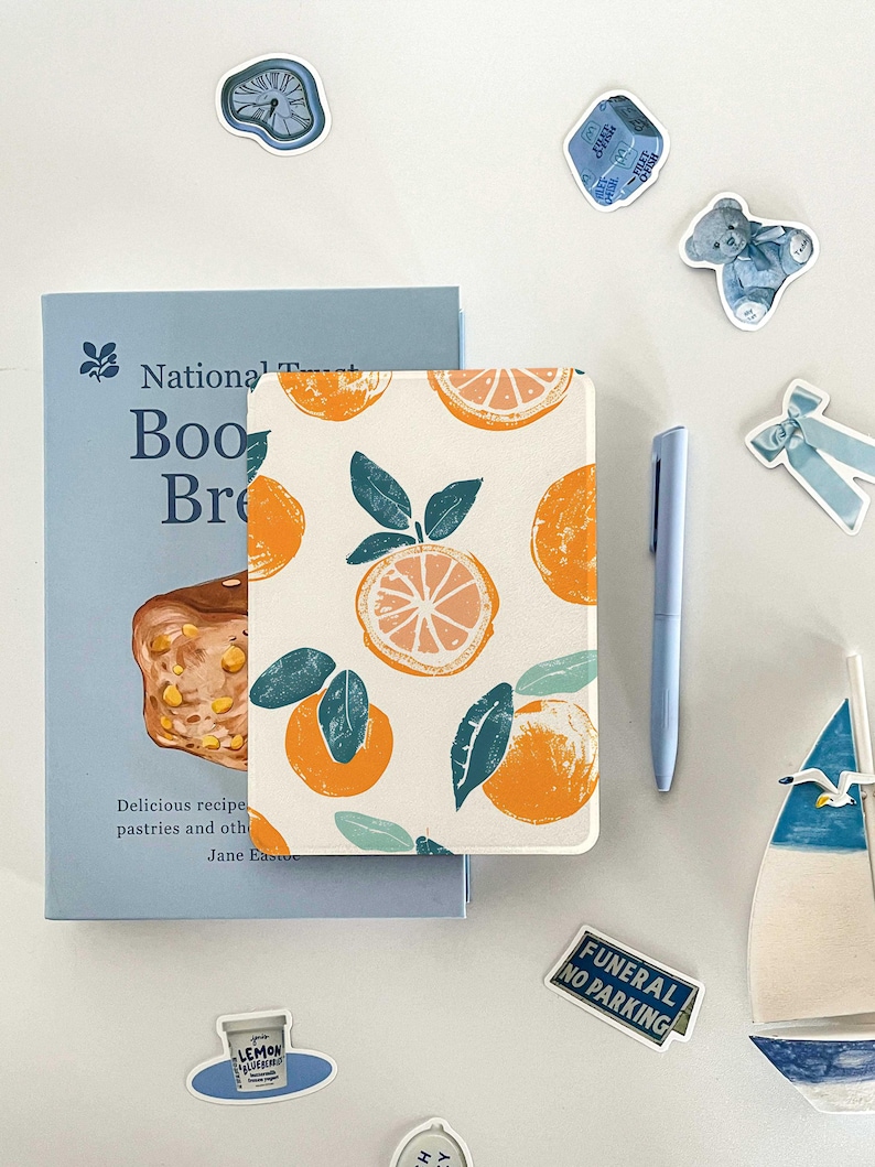 May include: A white notebook with a repeating pattern of orange slices and green leaves. The notebook is closed and lying on a white surface. There are several blue stickers around the notebook, including a clock, a teddy bear, a box, and a bow.