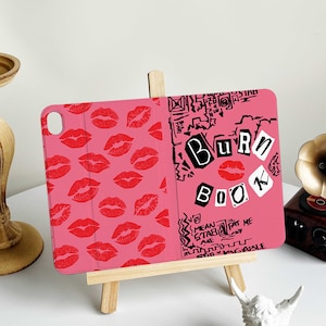May include: Pink tablet case with a design featuring red lip prints on the left side and the words "Burn Book" in a scrapbook style on the right. The case is propped up on a small wooden easel.