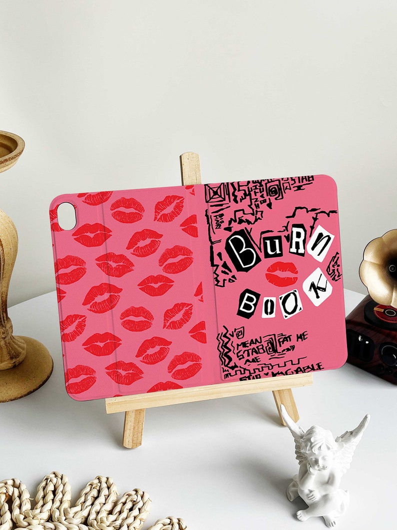 May include: Pink tablet case with a design featuring red lip prints on the left side and the words "Burn Book" in a scrapbook style on the right. The case is propped up on a small wooden easel.