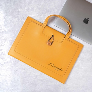 Könnte beinhalten: Senfgelbes Leder-Portfolio mit Griff und Holzknopfverschluss. Der Name "Maggie" ist in Schreibschrift auf der Vorderseite geschrieben. Ein Laptop ist im Hintergrund teilweise sichtbar.