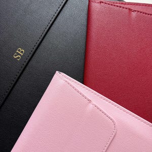 May include: An overhead shot of three leather notebooks in black, red, and pink. The black notebook has gold initials "SB" embossed on the cover. The notebooks have a stitched border and a smooth, matte finish.