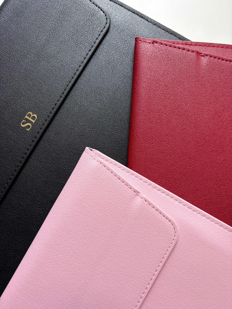 May include: An overhead shot of three leather notebooks in black, red, and pink. The black notebook has gold initials "SB" embossed on the cover. The notebooks have a stitched border and a smooth, matte finish.