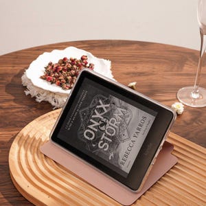 May include: A black Kindle e-reader in a clear protective case, resting on a light pink folding cover. The screen displays the book cover for Onyx Storm by Rebecca Yarros, featuring intricate line art and bold white text on a dark background.