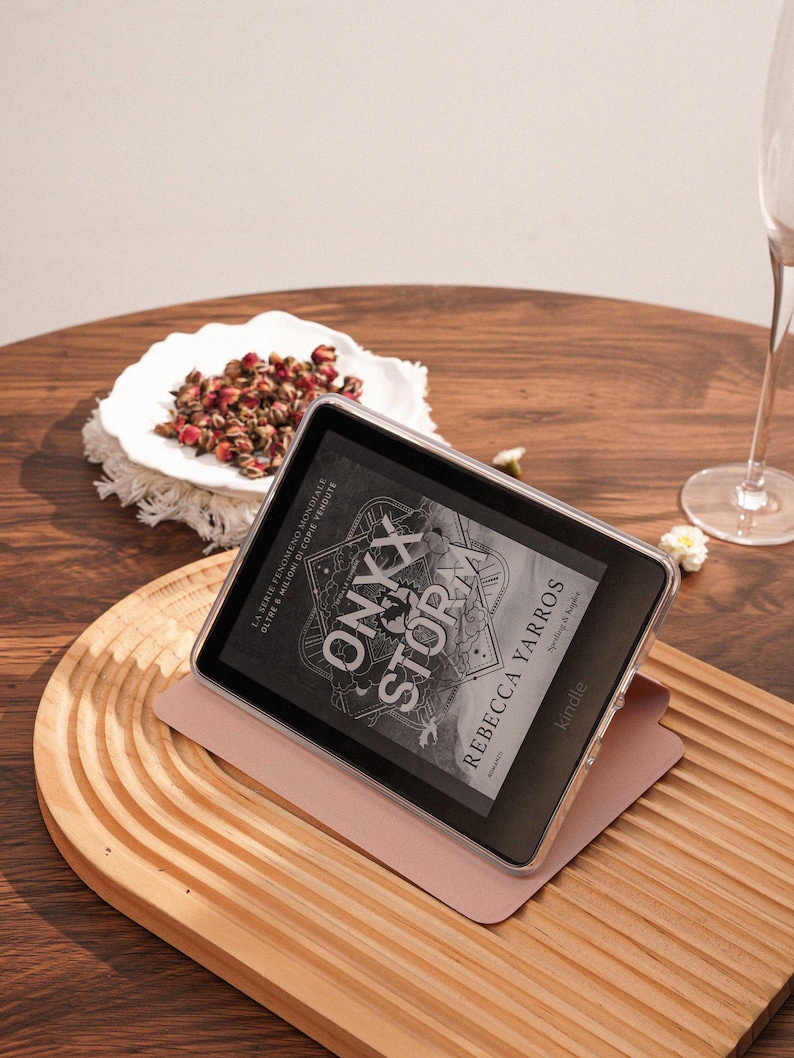 May include: A black Kindle e-reader in a clear protective case, resting on a light pink folding cover. The screen displays the book cover for Onyx Storm by Rebecca Yarros, featuring intricate line art and bold white text on a dark background.