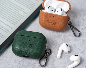 May include: Two personalized AirPod cases, one brown and one green, with the name "Maggie" engraved on them. The brown case is open, revealing white earbuds. Two white earbuds are also shown. Each case has a silver carabiner clip.