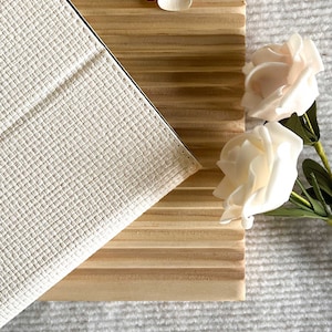 May include: A white leather notebook with a textured surface, lying on a wooden surface with a white flower and two white roses.