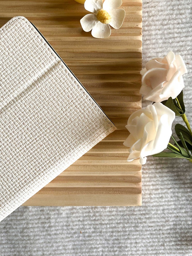 May include: A white leather notebook with a textured surface, lying on a wooden surface with a white flower and two white roses.