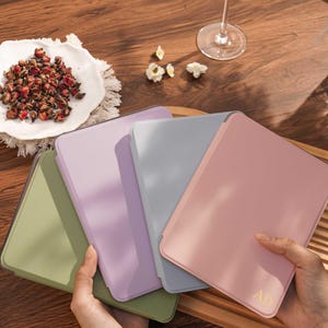 May include: Four rectangular tablet cases in olive green, lavender, light gray, and dusty pink. The pink case features the gold embossed letters AD in the bottom right corner. Each case has a smooth, solid-colored finish and rounded corners.