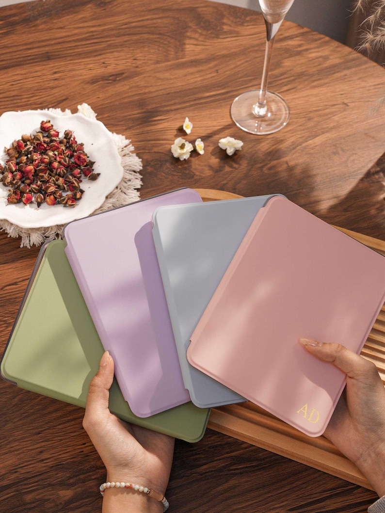 May include: Four rectangular tablet cases in olive green, lavender, light gray, and dusty pink. The pink case features the gold embossed letters AD in the bottom right corner. Each case has a smooth, solid-colored finish and rounded corners.