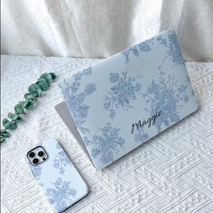 May include: A light blue laptop cover and a matching smartphone case, both featuring a dark blue floral pattern. The laptop cover is personalized with the name Maggie in a black cursive script. The items are displayed together as a set.