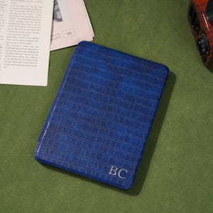 May include: A blue, rectangular case with a crocodile skin texture. The initials "BC" are embossed in the lower right corner. The case is on a green surface, with a newspaper and a radio in the background.