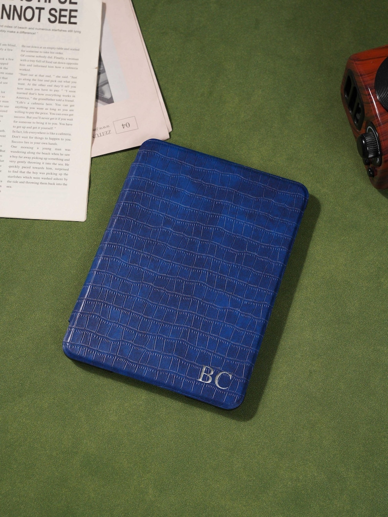 May include: A blue, rectangular case with a crocodile skin texture. The initials "BC" are embossed in the lower right corner. The case is on a green surface, with a newspaper and a radio in the background.