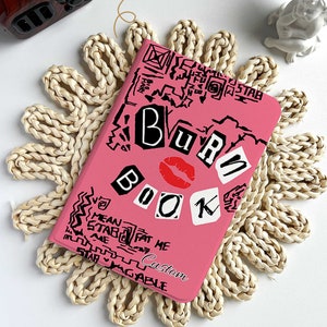 May include: Pink notebook with the words "BURN BOOK" in a collage style, featuring a red lip graphic. The notebook is surrounded by a woven placemat. The text "Custom" is also visible on the notebook.