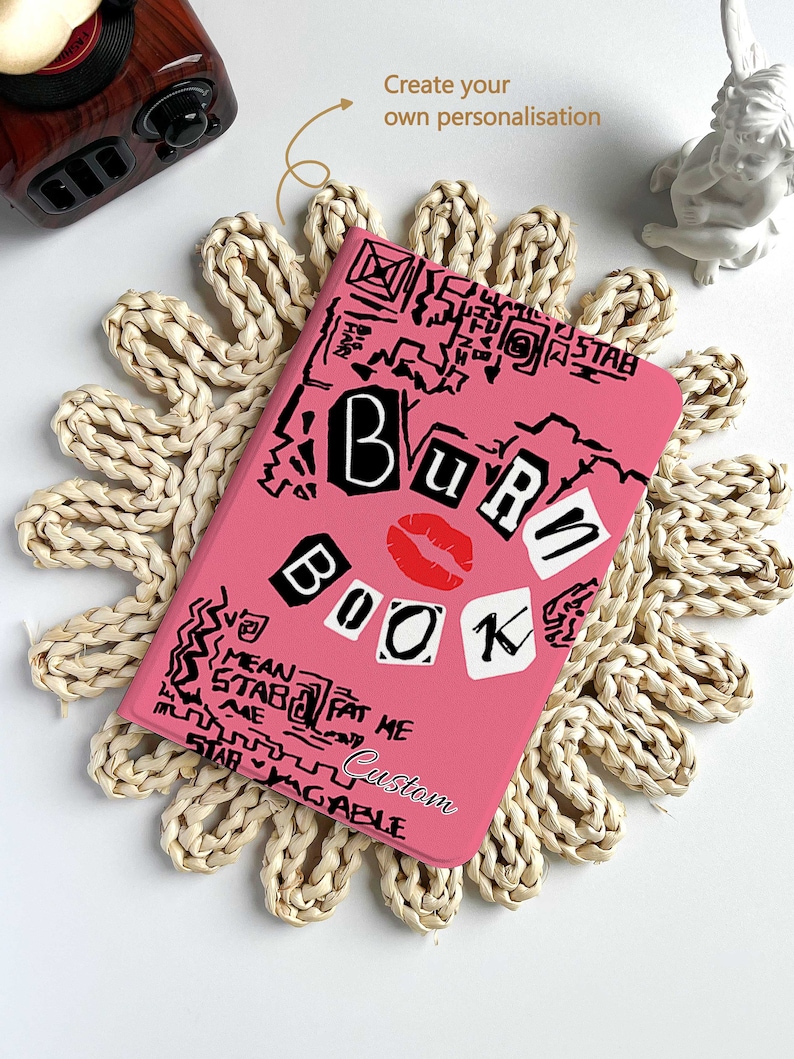 May include: Pink notebook with the words "BURN BOOK" in a collage style, featuring a red lip graphic. The notebook is surrounded by a woven placemat. The text "Custom" is also visible on the notebook.