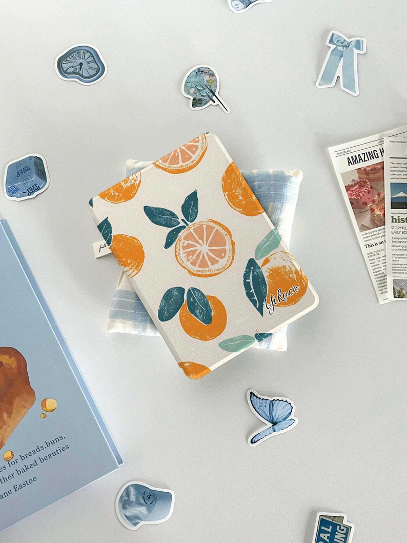 May include: A white notebook cover with a repeating pattern of orange citrus fruit and green leaves. The notebook cover has the name "Yikoco" written in black cursive on the bottom right corner.