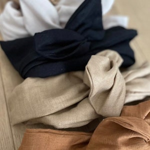 May include: A stack of four fabric headbands in different colours, including black, beige, and brown. The headbands are made of a soft, textured fabric and have a knotted bow design.