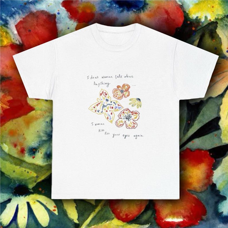 Puede incluir: Camiseta blanca con un dise&ntilde;o de mariposa y flores y el texto "I don't wanna talk about anything... I wanna kiss your eyes again." El dise&ntilde;o incluye una mariposa y flores en tonos amarillos, naranjas y azules.
