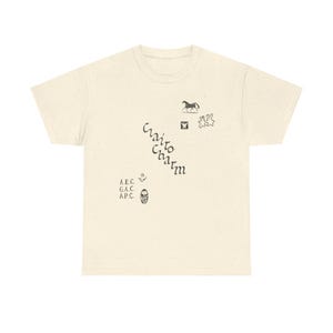 May include: Cream-colored t-shirt with a speckled texture. The shirt features various small black illustrations, including a horse, a butterfly, and abstract shapes. The words "Cairo Charm" are printed diagonally, along with the initials "A.E.C., G.A.C., A.P.C."