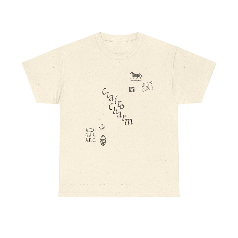 May include: Cream-colored t-shirt with a speckled texture. The shirt features various small black illustrations, including a horse, a butterfly, and abstract shapes. The words "Cairo Charm" are printed diagonally, along with the initials "A.E.C., G.A.C., A.P.C."