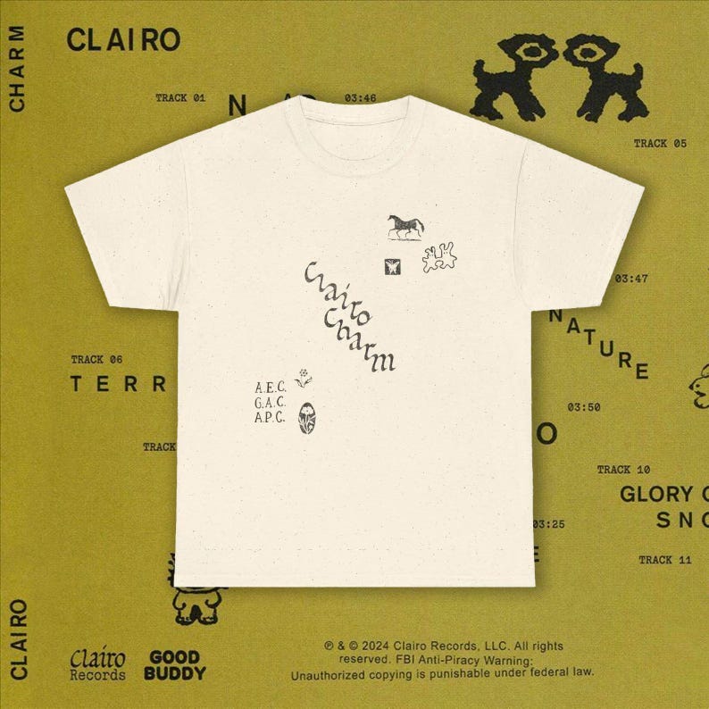 May include: Cream-colored t-shirt with various small black graphics and the text "Clairo Charm" in a handwritten font. The shirt is set against a mustard-colored background with additional text and illustrations, including "Clairo Records" and track listings.