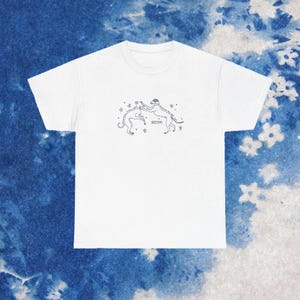 May include: White t-shirt featuring a minimalist line drawing of two dogs playfully interacting, surrounded by stars. The design is in black and is centered on the shirt. The shirt is made of a soft material.