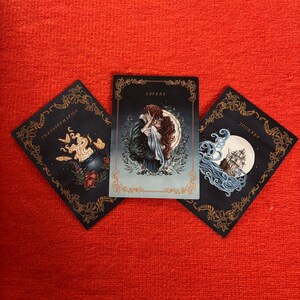 May include: Three tarot cards on a red surface. The center card reads "Lovers" and depicts a couple embracing. The other cards read "Transformation" and "Journey" and feature illustrations with gold floral borders and dark backgrounds.