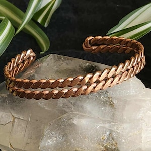 May include: A copper-colored, braided cuff bracelet. The bracelet is resting on a clear, crystalline rock. Green and white striped leaves are in the background. The bracelet has a twisted rope design.