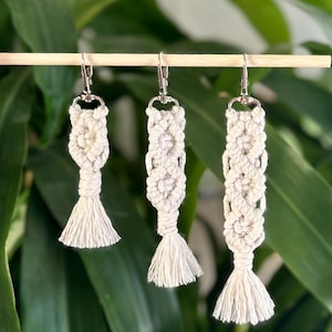 May include: Three handmade macrame earrings with silver-colored hooks. The earrings are off-white, with a knotted design and a tassel at the bottom. The background is a lush green plant.
