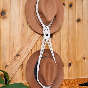 May include: A macrame hat holder with two brown hats hanging on a wooden door. The white macrame has a wooden ring at the top and decorative knots with dark brown beads. The hats are a light brown color.