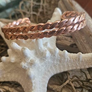 May include: A copper-colored cuff bracelet with a twisted rope design. The bracelet is displayed on a white starfish, with driftwood and fishing net in the background. The bracelet's design is visible.
