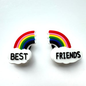 May include: Two colorful rainbow charms with the words "BEST" and "FRIENDS" on white cloud-shaped bases. The rainbows feature red, pink, yellow, green, blue, and black stripes. These charms are likely for shoes or accessories.
