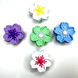 Könnte beinhalten: Eine Sammlung von sechs blütenförmigen Verzierungen in verschiedenen Farben. Die Blumen sind weiß, lila, grün und blau, mit gelben, weißen, roten und violetten Zentren. Ideal für Bastelarbeiten und DIY-Projekte.