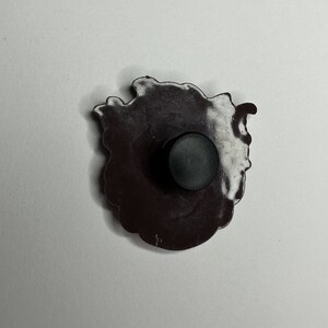 Puede incluir: Un pin de esmalte blanco y negro con un dise&ntilde;o detallado de una flor. El pin tiene un respaldo de goma negro.