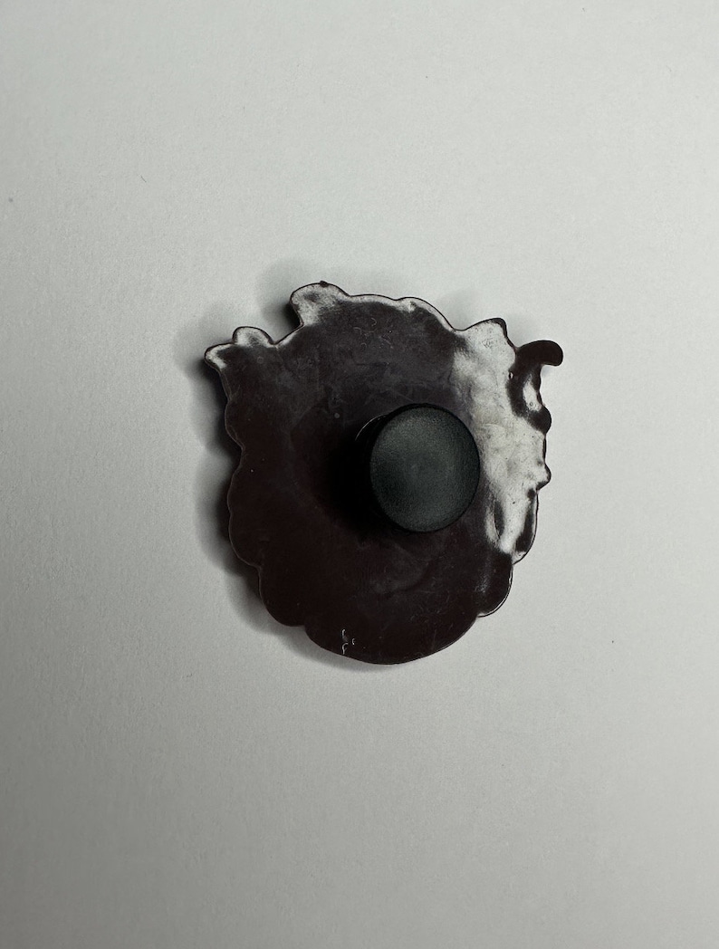Puede incluir: Un pin de esmalte blanco y negro con un dise&ntilde;o detallado de una flor. El pin tiene un respaldo de goma negro.